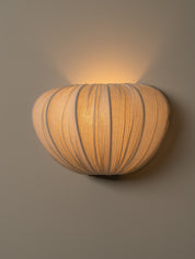 A warm, ribbed wall sconce light fixture with a rounded, pumpkin-like shape, mounted on a neutral gray wall and casting a soft glow upward.
