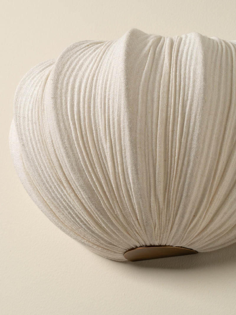 A close-up of a pleated fabric wall sconce in a warm off-white or cream color, showing the detailed texture of the folds and a small metallic gold accent at the bottom, set against a solid beige background.