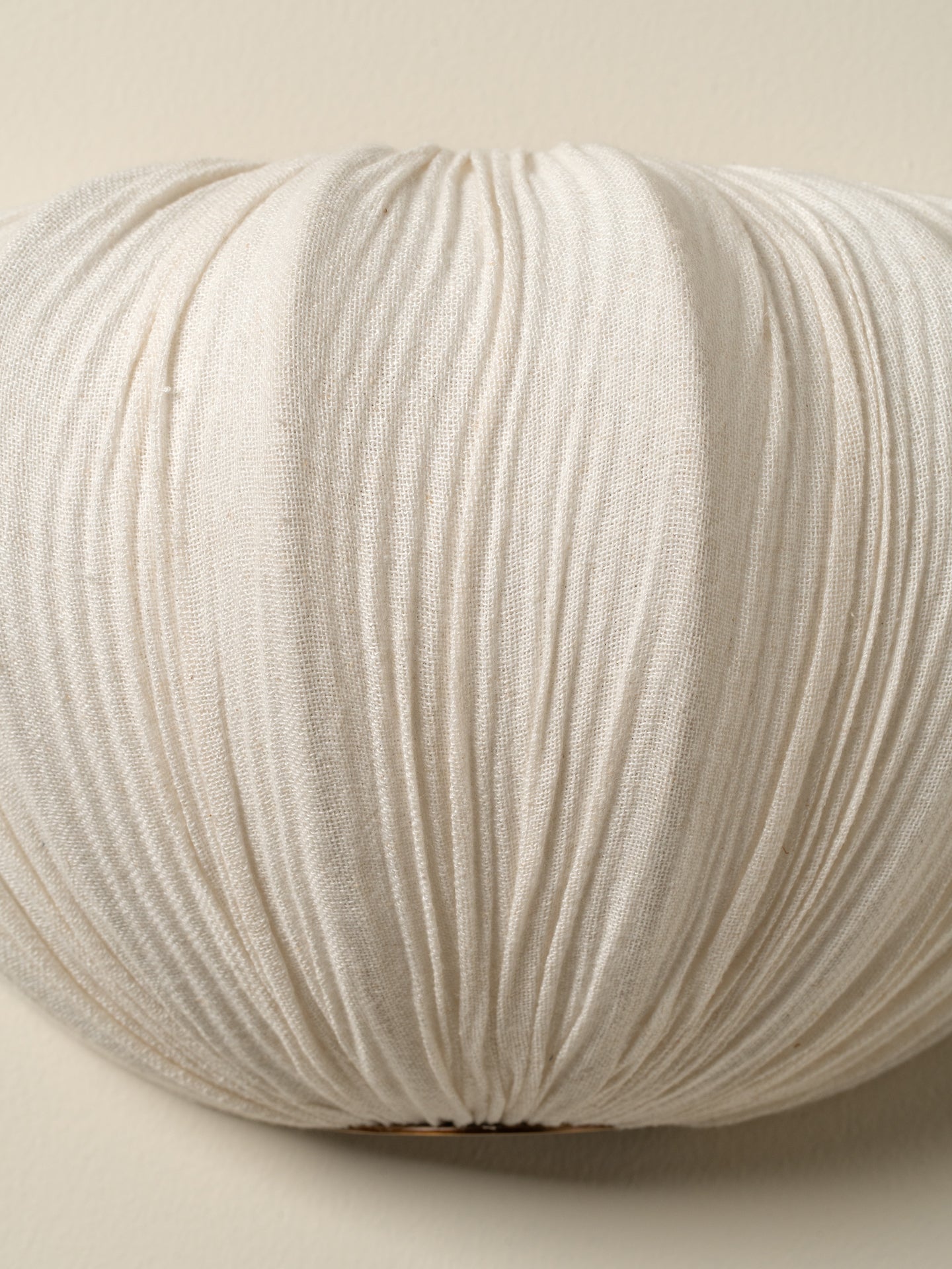 A close-up of a round, pleated decorative pillow in a warm off-white or cream color, showing the textured fabric and gathered pleats against a neutral beige background.