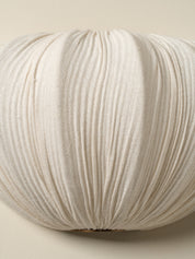 A close-up of a round, pleated decorative pillow in a warm off-white or cream color, showing the textured fabric and gathered pleats against a neutral beige background.