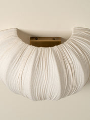 A close-up of a pleated, cloud-like white wall sconce with a brass metal mounting bracket, mounted on a warm beige wall.