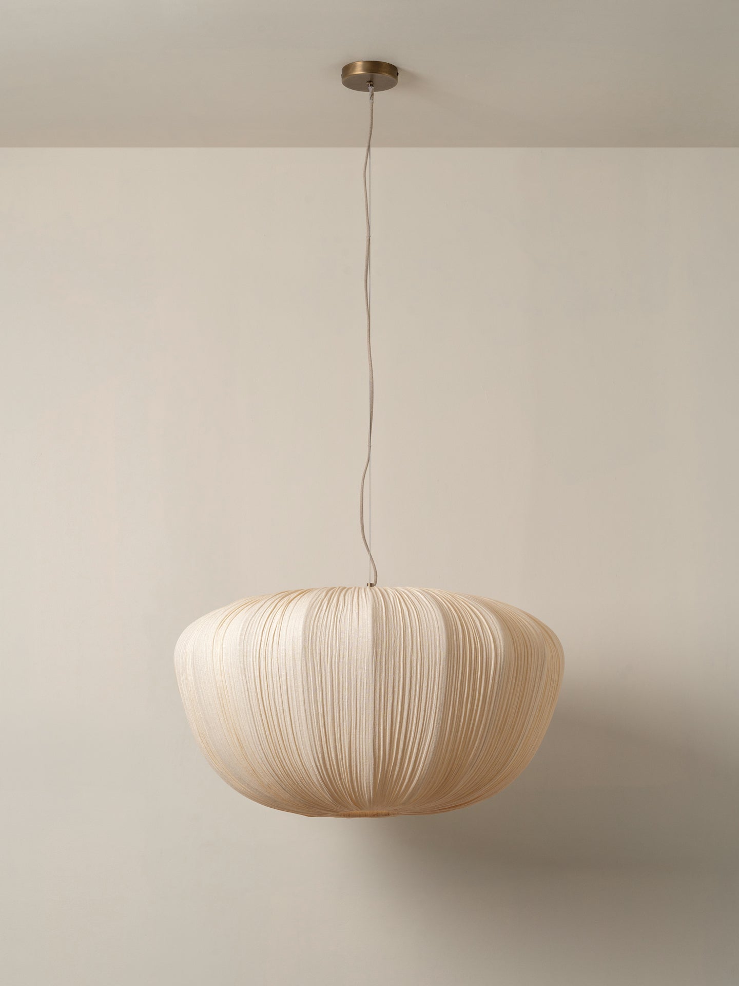 A modern pleated paper pendant light in a warm cream color, suspended from a brass ceiling canopy against a minimalist beige wall.