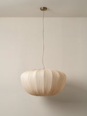 A modern pleated paper pendant light in a warm cream color, suspended from a brass ceiling canopy against a minimalist beige wall.