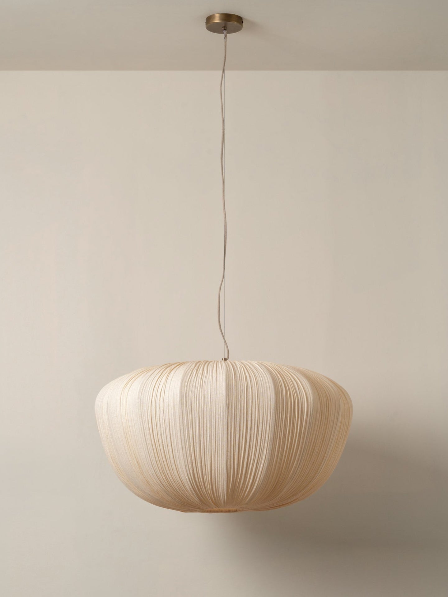A modern pleated paper pendant light in a warm cream color, suspended from a brass ceiling canopy against a minimalist beige wall.