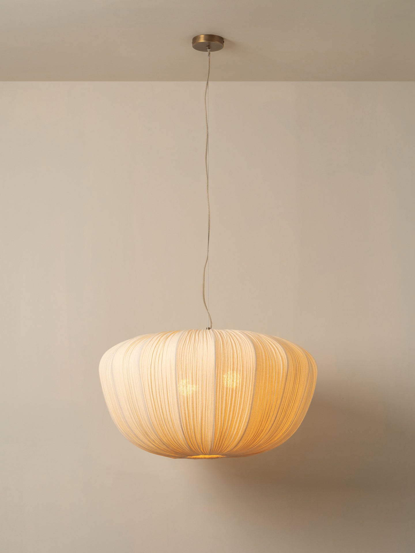 A modern pendant light with a pleated, ribbed shade made of natural beige material, suspended by a thin cord from a brass ceiling canopy against a neutral beige wall.