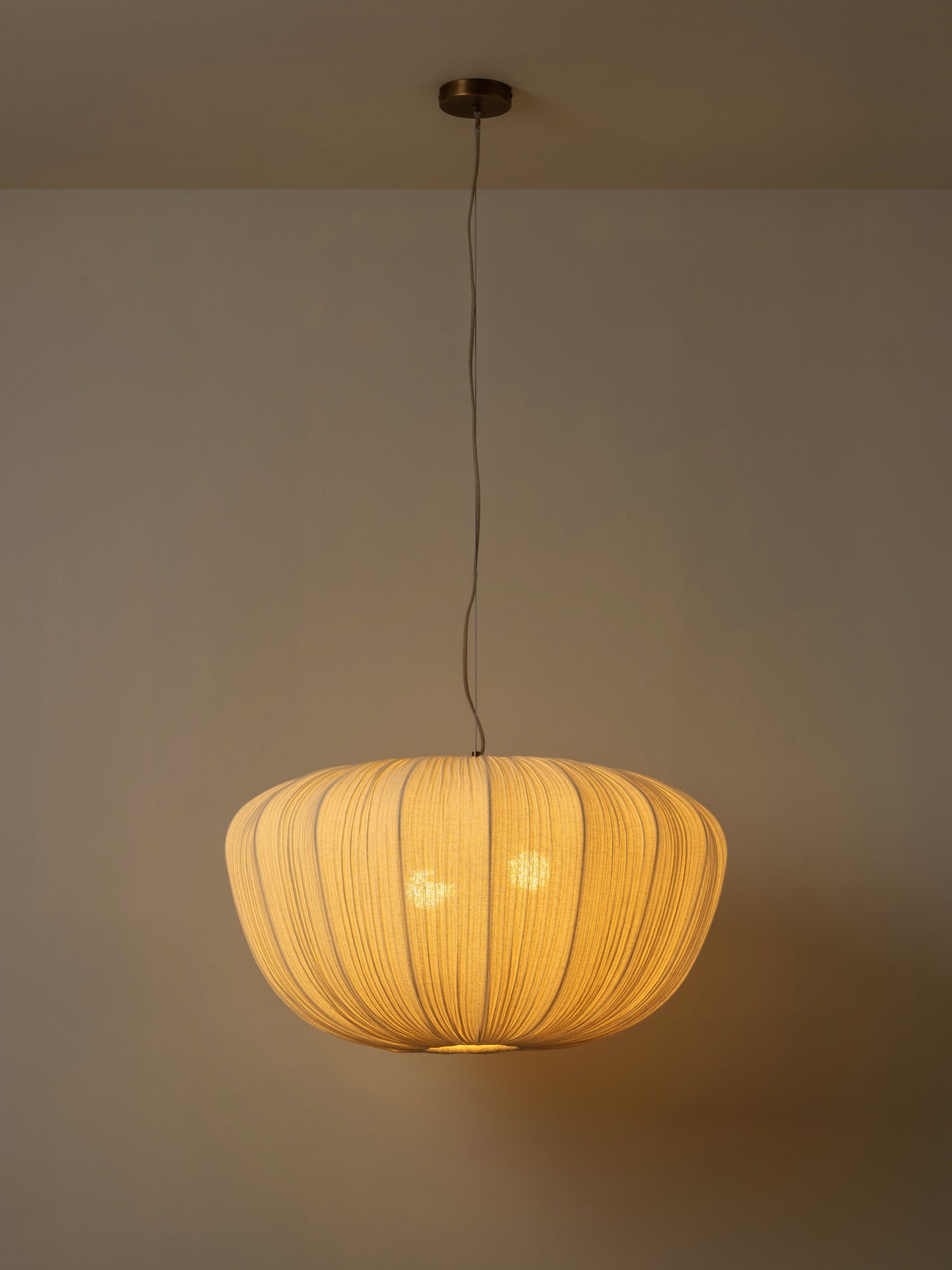 A modern pendant light with a ribbed, pleated shade made of warm beige fabric, suspended from a ceiling by a thin black cord. The light is turned on, casting a warm yellow glow against a neutral beige wall.