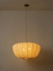 A modern pendant light with a ribbed, pleated shade made of warm beige fabric, suspended from a ceiling by a thin black cord. The light is turned on, casting a warm yellow glow against a neutral beige wall.