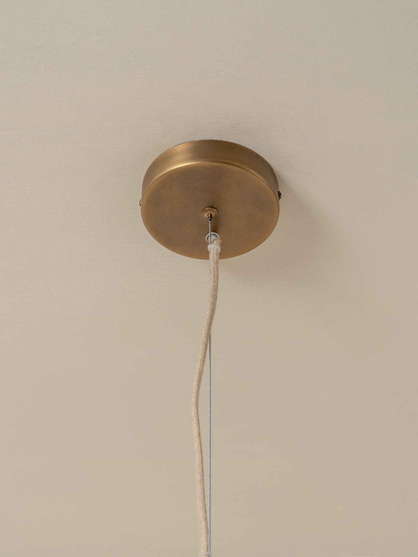 A close-up of a ceiling-mounted pendant light fixture showing a round brass ceiling canopy with a white cord hanging down against a plain off-white ceiling.