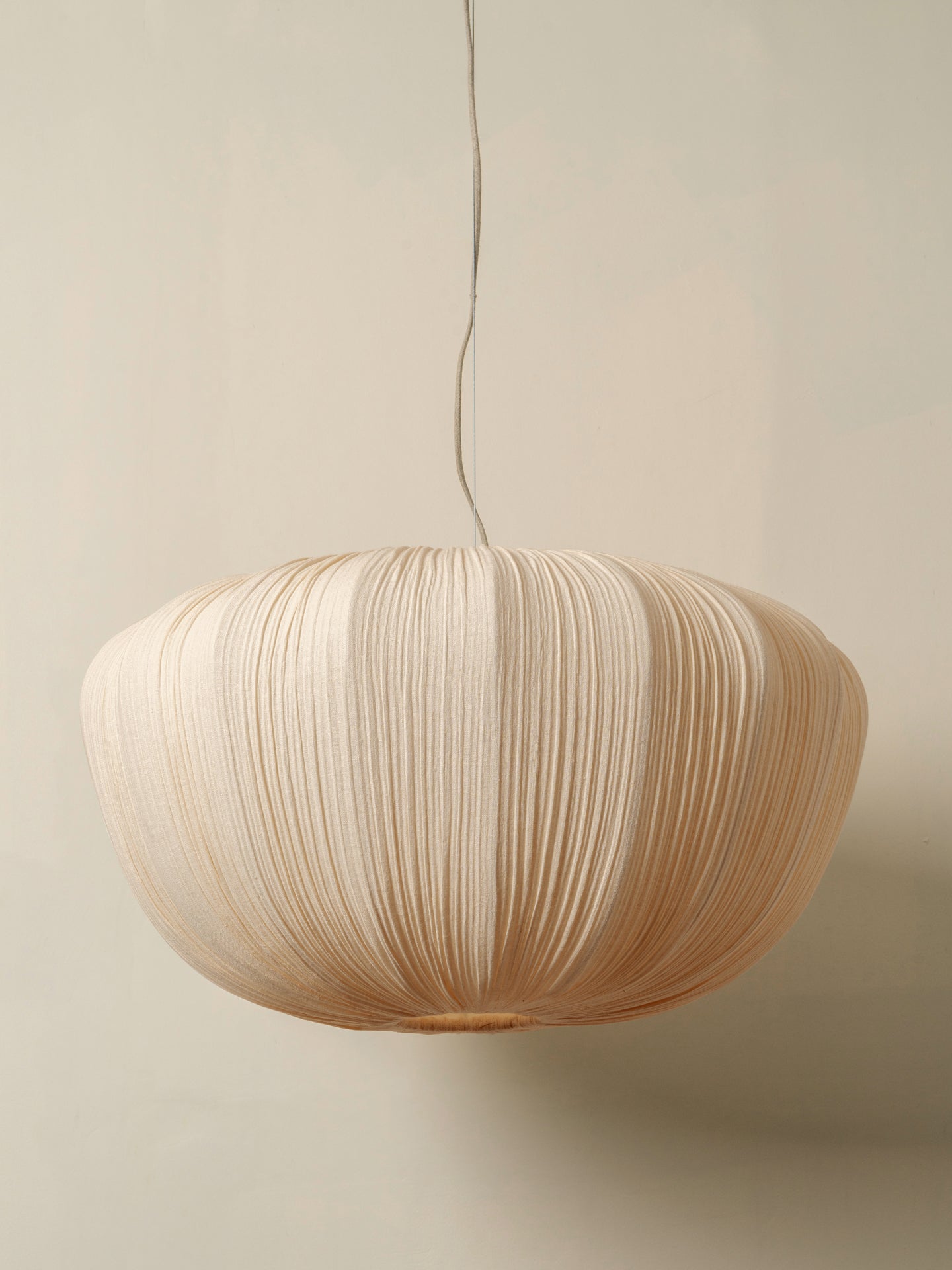 A modern pendant light with a large, rounded shade made of pleated natural paper or fabric, suspended by a thin cord against a warm beige wall.