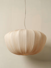 A modern pendant light with a large, rounded shade made of pleated natural paper or fabric, suspended by a thin cord against a warm beige wall.