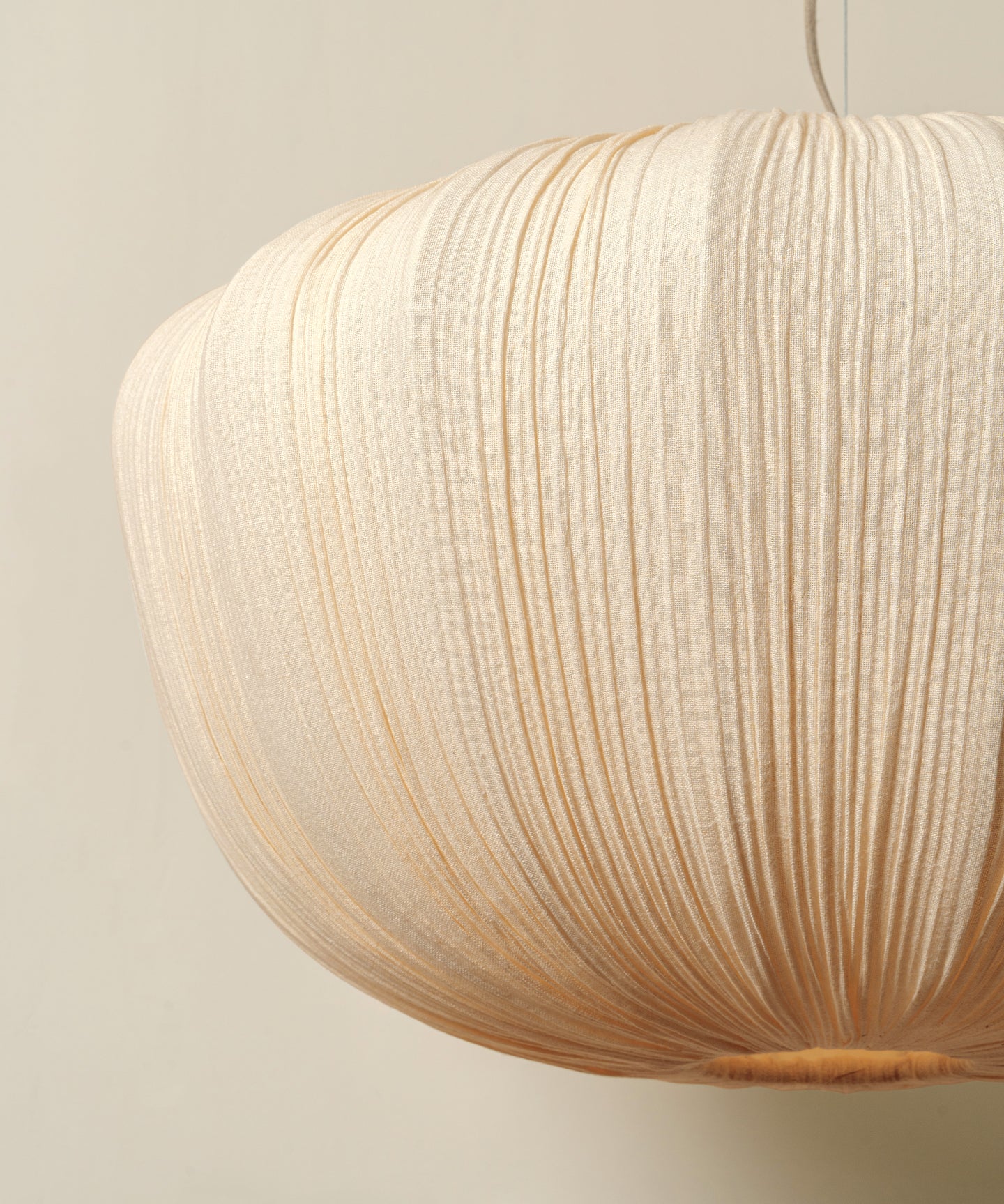 A close-up of a pleated paper pendant light in a warm cream or natural beige color, hanging against a solid light beige wall.