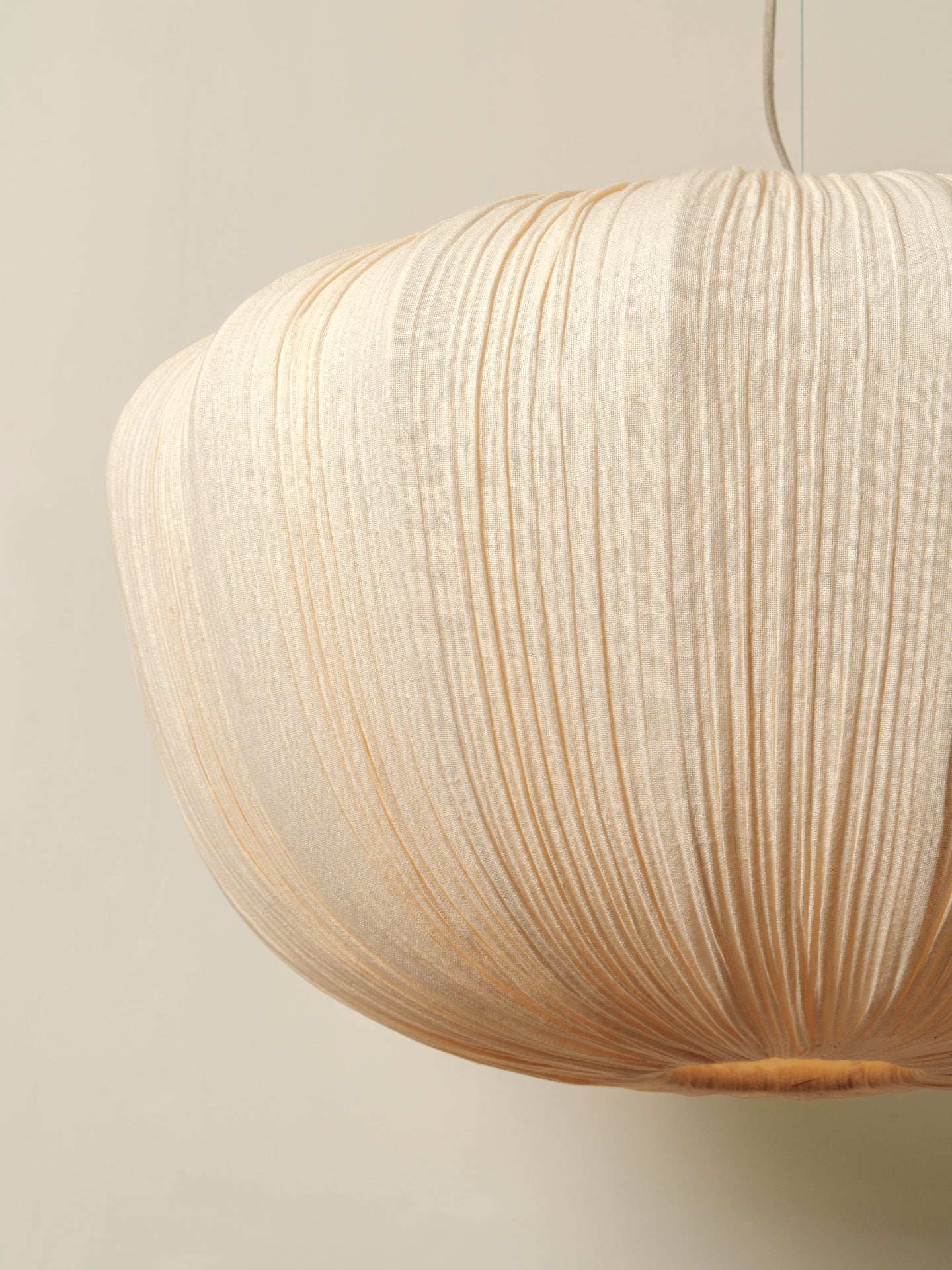 A close-up of a pleated paper pendant light in a warm cream or natural beige color, hanging against a solid light beige wall.