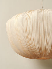 A close-up of a pleated paper pendant light in a warm cream or natural beige color, hanging against a solid light beige wall.