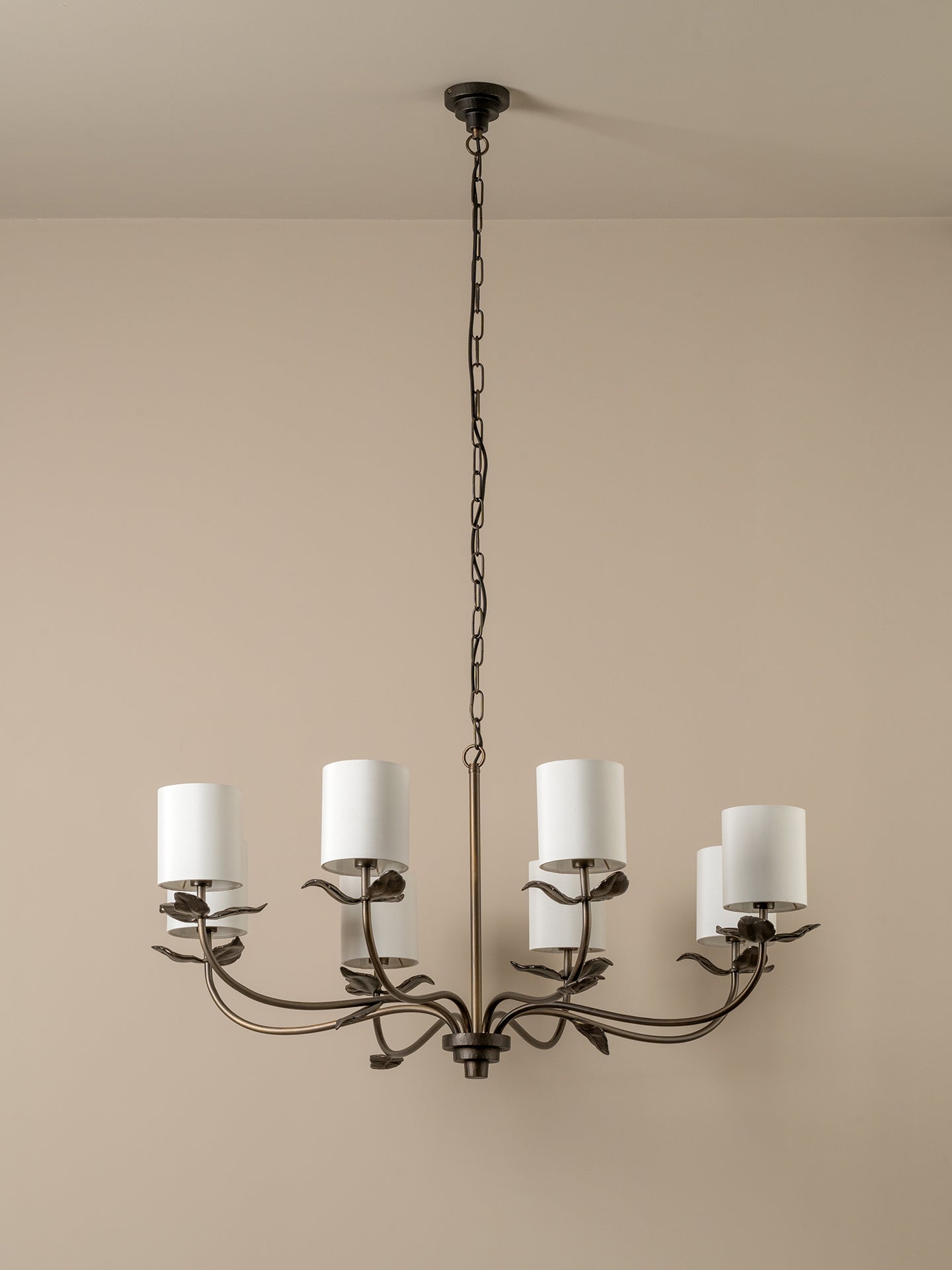 A six-light chandelier with a dark metal leaf-style frame and white cylindrical drum shades, hanging from a chain against a beige wall.