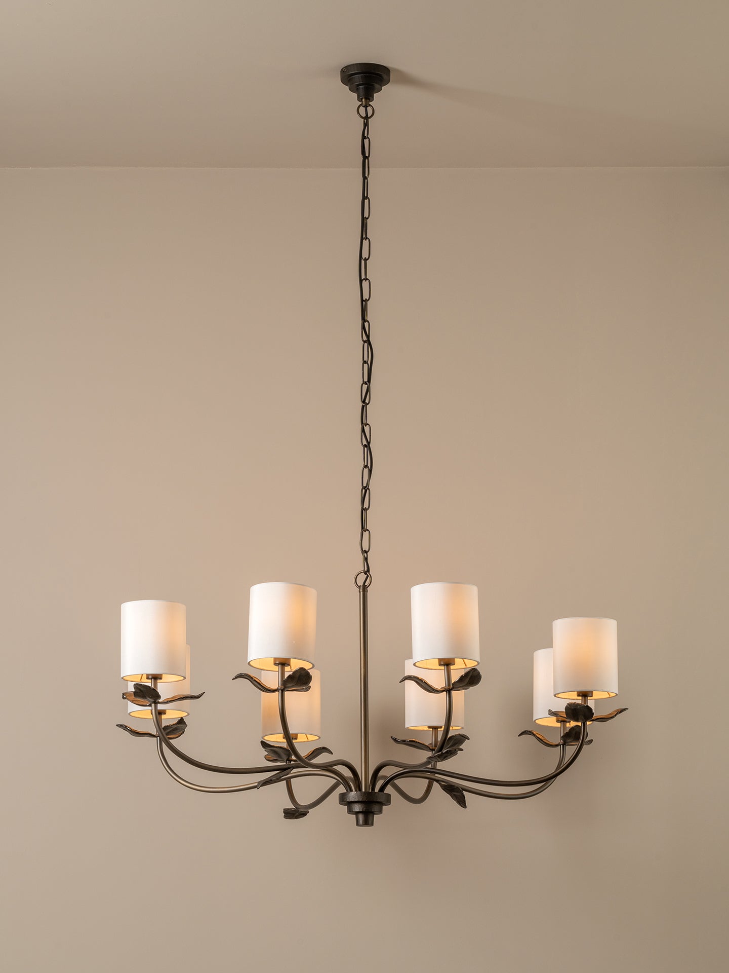 A six-light chandelier with a dark metal frame and white cylindrical drum shades, suspended by a chain from a ceiling against a plain beige wall.