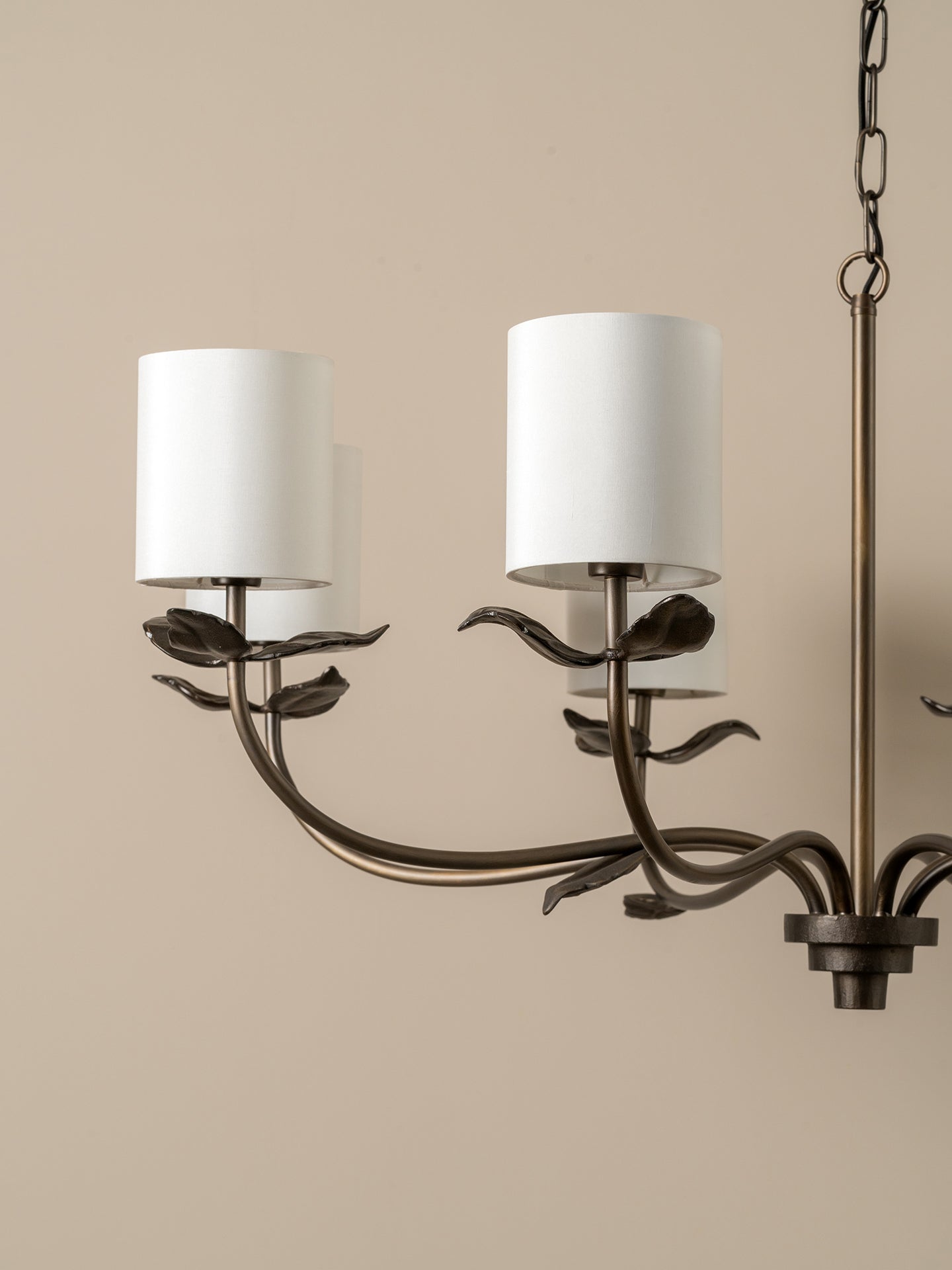 A close-up of a multi-light chandelier with a dark bronze metal frame designed to look like climbing vines or leaves, featuring two white drum shades and a hanging chain against a solid beige background.