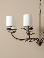 A close-up of a multi-light chandelier with a dark bronze metal frame designed to look like climbing vines or leaves, featuring two white drum shades and a hanging chain against a solid beige background.