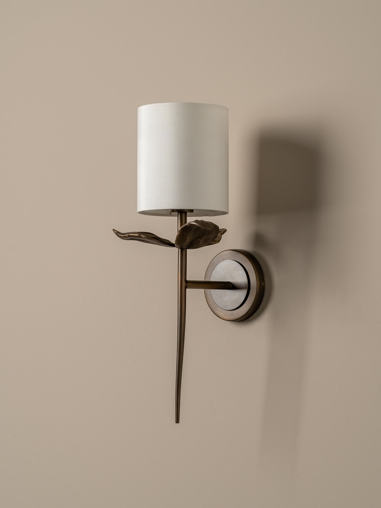 A wall-mounted sconce with a white cylindrical shade, a dark bronze metal stem and leaf detail, and a round backplate on a warm beige wall.