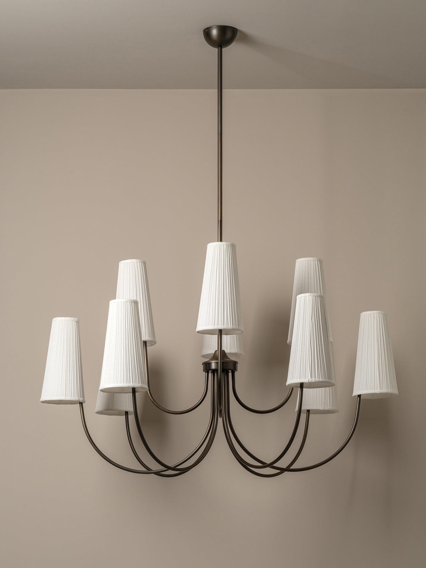 A modern nine-light chandelier with a dark metal frame and pleated white drum shades, shown hanging against a neutral beige wall.