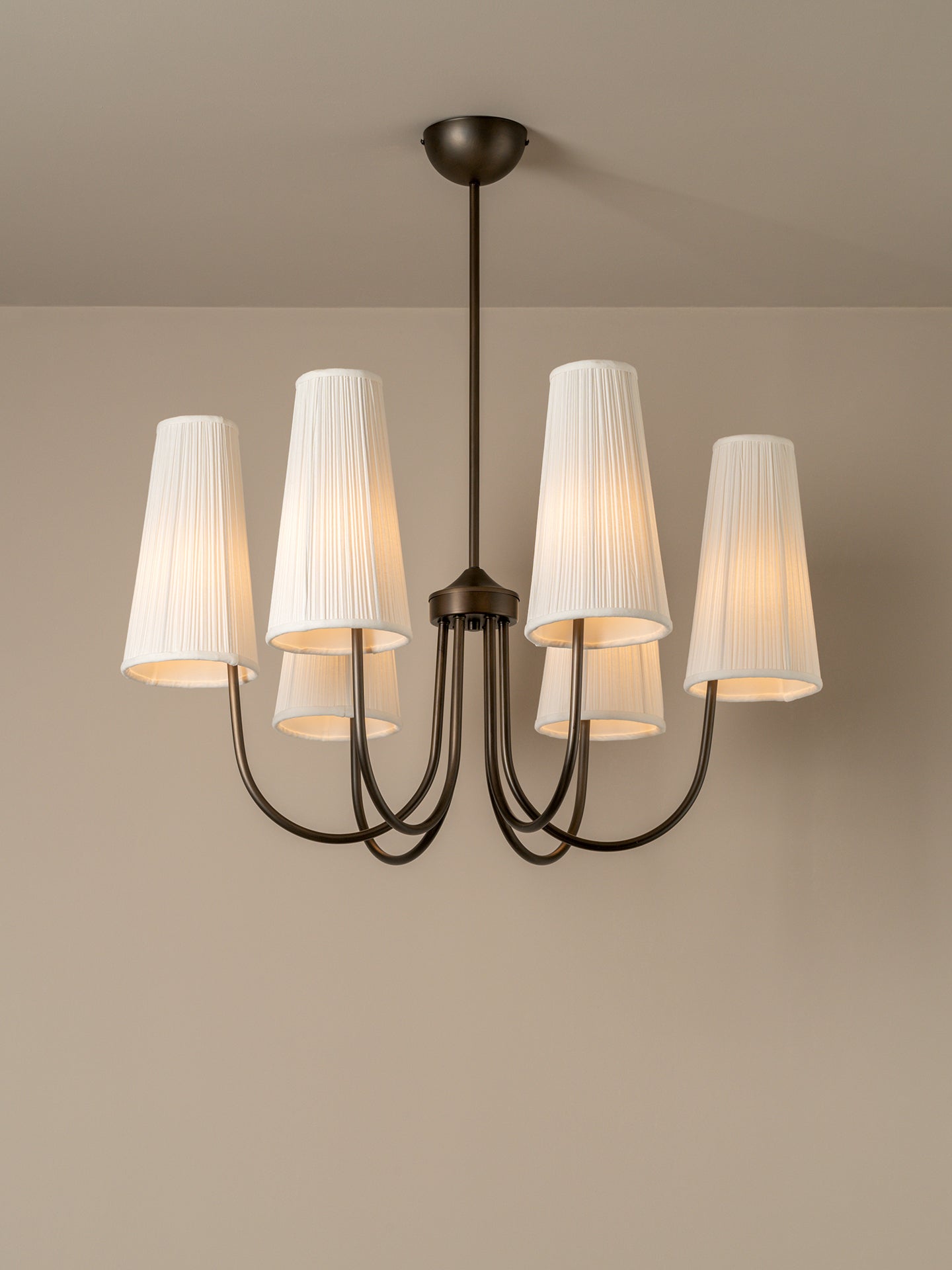 A six-light chandelier with a dark bronze metal frame and white pleated fabric shades, shown illuminated against a neutral beige wall and ceiling.