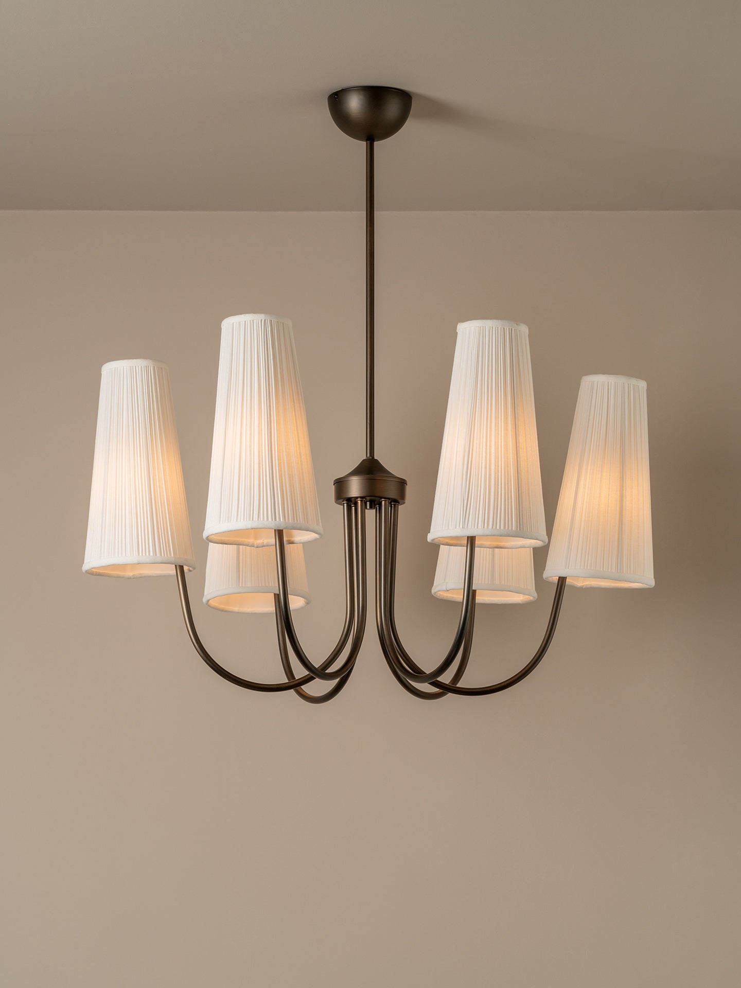 A five-light chandelier with a dark bronze metal frame and white pleated fabric shades, shown illuminated against a neutral beige wall and ceiling.