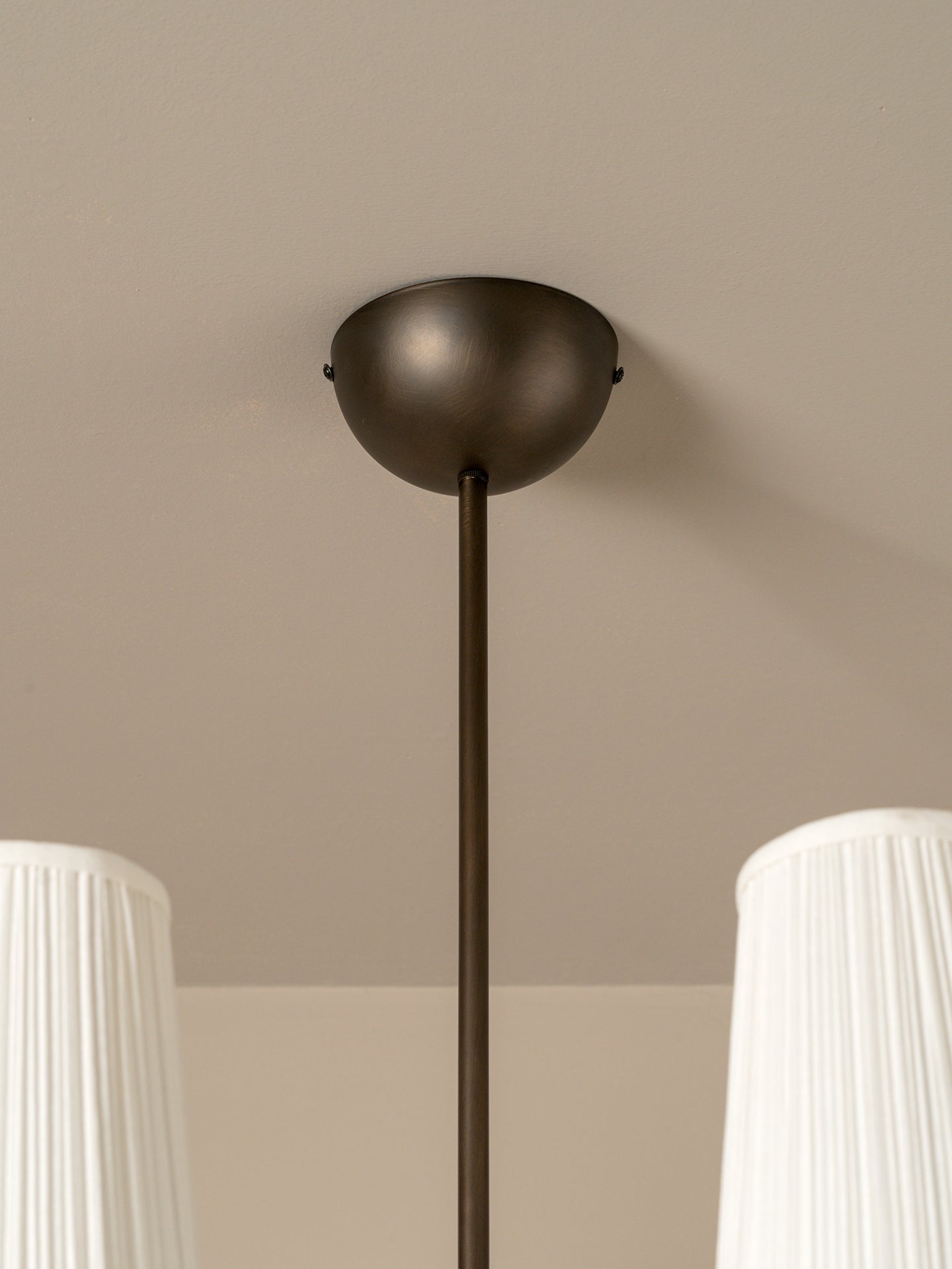 A close-up of a modern ceiling light fixture with a matte dark bronze dome canopy and a matching vertical rod, shown installed on a beige ceiling with white pleated lampshades partially visible at the bottom corners.