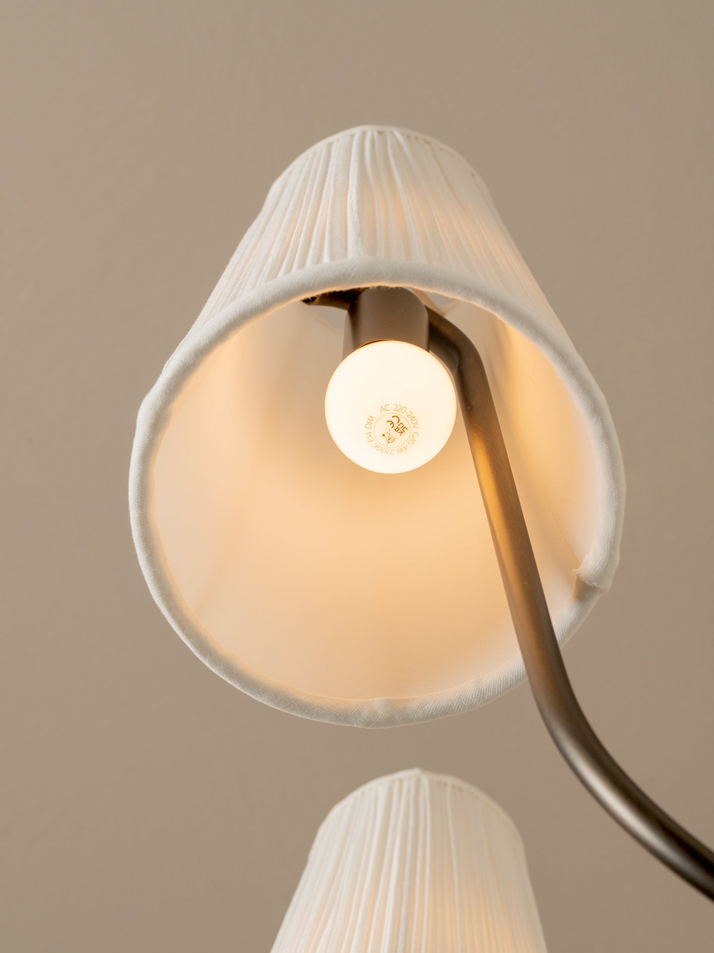 A close-up of a modern floor lamp with a pleated white fabric shade and a curved dark metal arm, shown lit against a warm beige background.