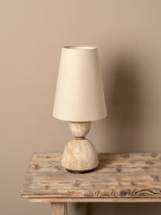 A table lamp with a tapered off-white fabric shade and a textured, rounded beige stone-like base, displayed on a rustic wooden table against a solid beige wall.