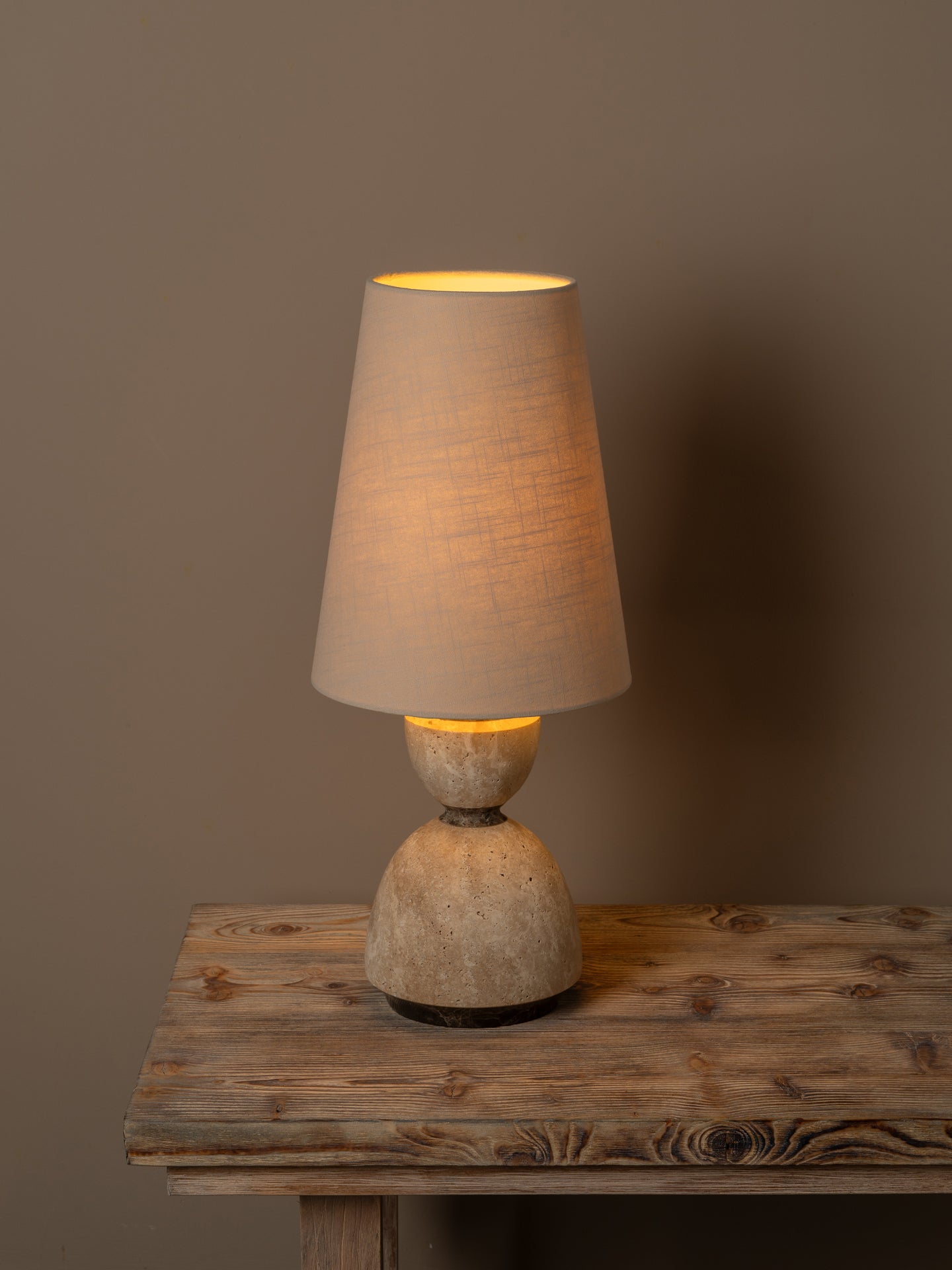 A rustic table lamp with a textured beige fabric drum shade and a carved stone-like base, illuminated with a warm yellow glow, set on a weathered wooden table against a solid taupe background.
