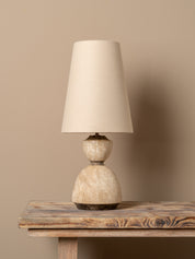 A table lamp with a tapered off-white fabric shade and a textured, double-bulb stone base in beige and cream tones, resting on a rustic wooden table against a solid beige wall.