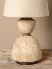 A table lamp with a textured beige stone base and a matching fabric drum shade, set on a rustic wooden surface against a neutral beige background.