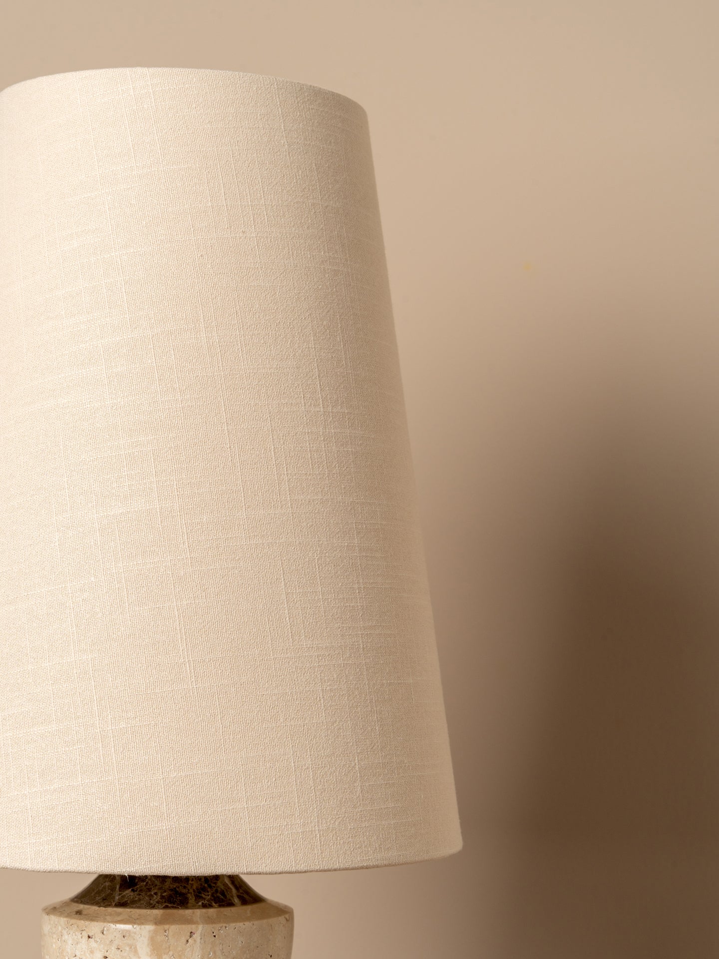 A close-up of a table lamp featuring a large, tapered off-white linen shade and a textured stone base against a warm beige background.