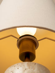 Close-up of a modern pendant light with a textured off-white fabric drum shade, a warm brass cylindrical socket, and a white speckled stone or concrete base, illuminated from within.