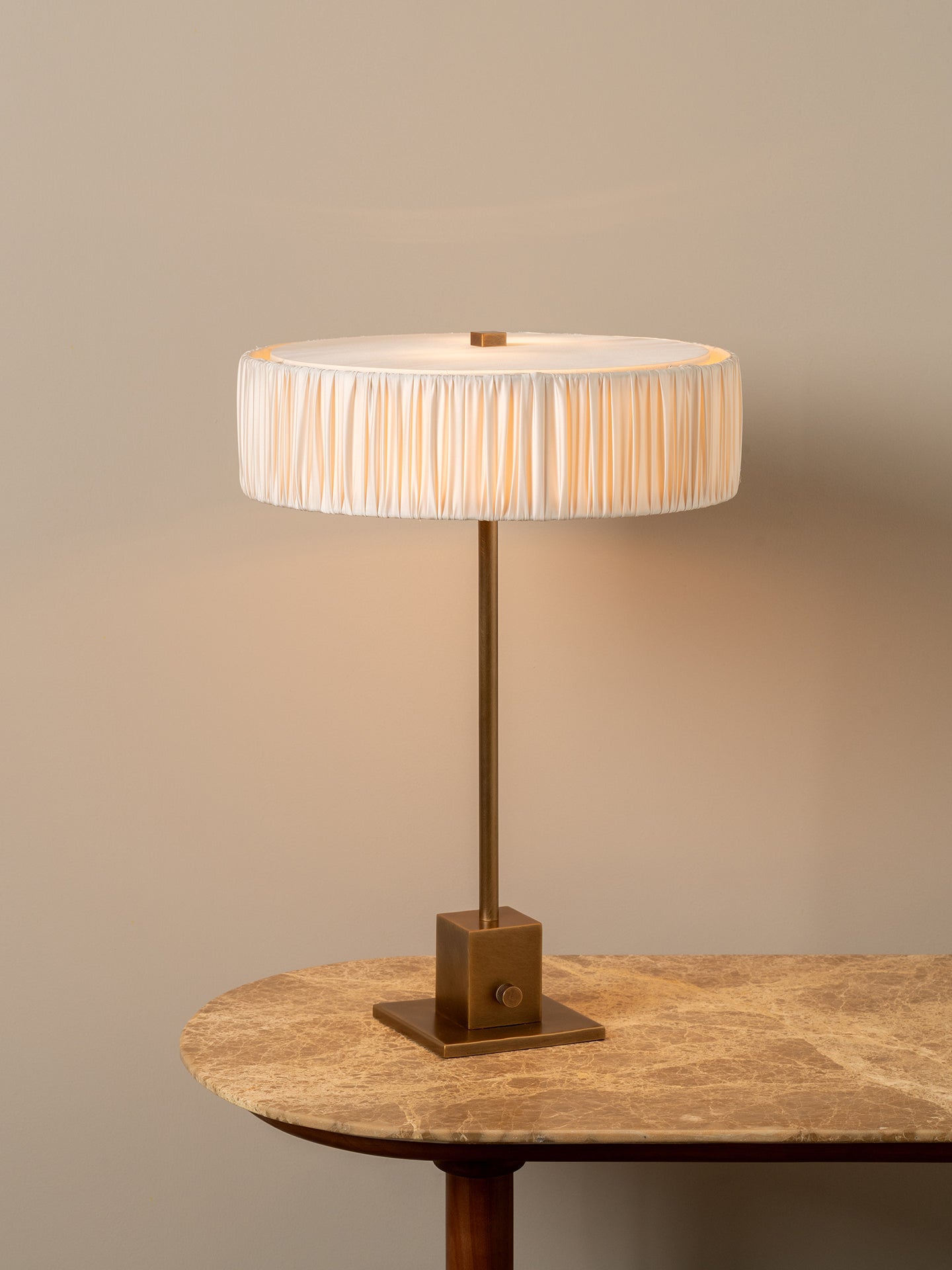 A table lamp with a pleated white drum shade and a square brass base, shown lit on a stone-topped table against a warm beige wall.