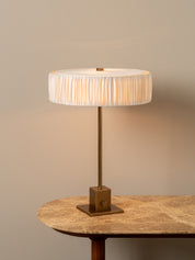 A table lamp with a pleated white drum shade and a square brass base, shown lit on a stone-topped table against a warm beige wall.