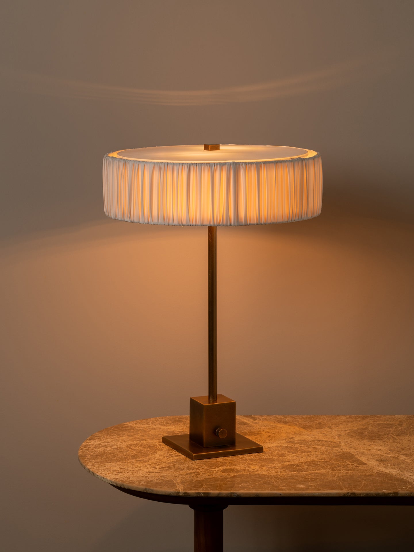 A table lamp with a pleated white drum shade and a square brass base, illuminated with a warm golden glow, set on a stone-topped table against a neutral background.