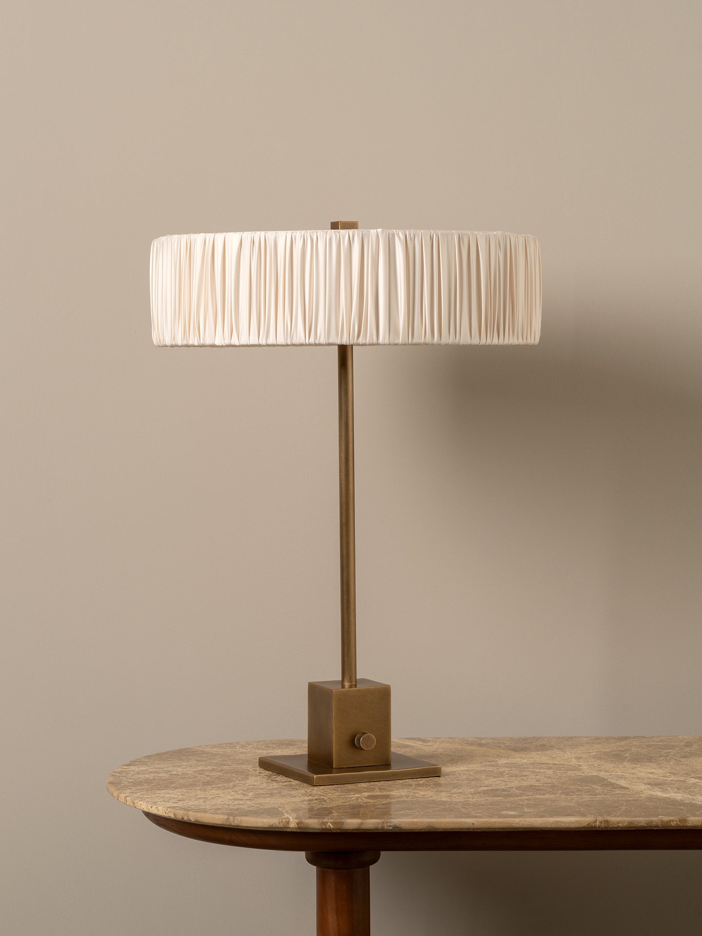A table lamp with a pleated cream fabric drum shade and a bronze-finished metal base, shown on a stone-topped table against a warm beige wall.