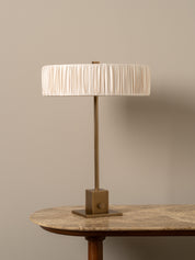 A table lamp with a pleated cream fabric drum shade and a bronze-finished metal base, shown on a stone-topped table against a warm beige wall.