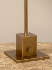 A close-up of a bronze-finished table lamp base with a square pedestal, flat square base plate, and vertical stem, shown on a textured beige surface against a neutral beige background.