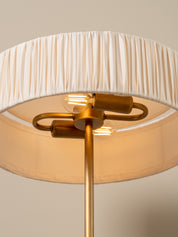 A close-up of a modern floor lamp featuring a pleated drum shade in warm off-white, a brushed brass metal stand, and two visible glowing Edison-style light bulbs.