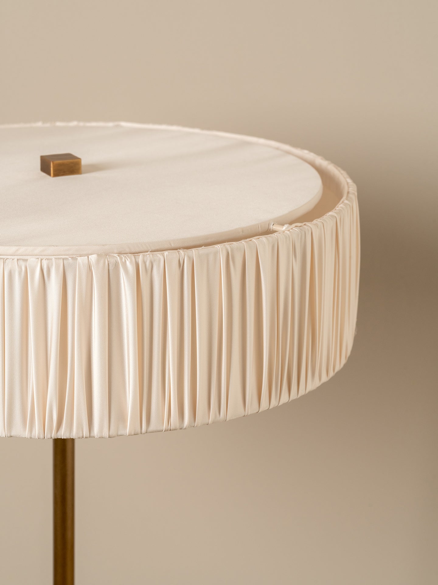 Close-up of a round table with a pleated cream fabric skirt and a small metallic gold square accent on the tabletop, set against a warm beige background.