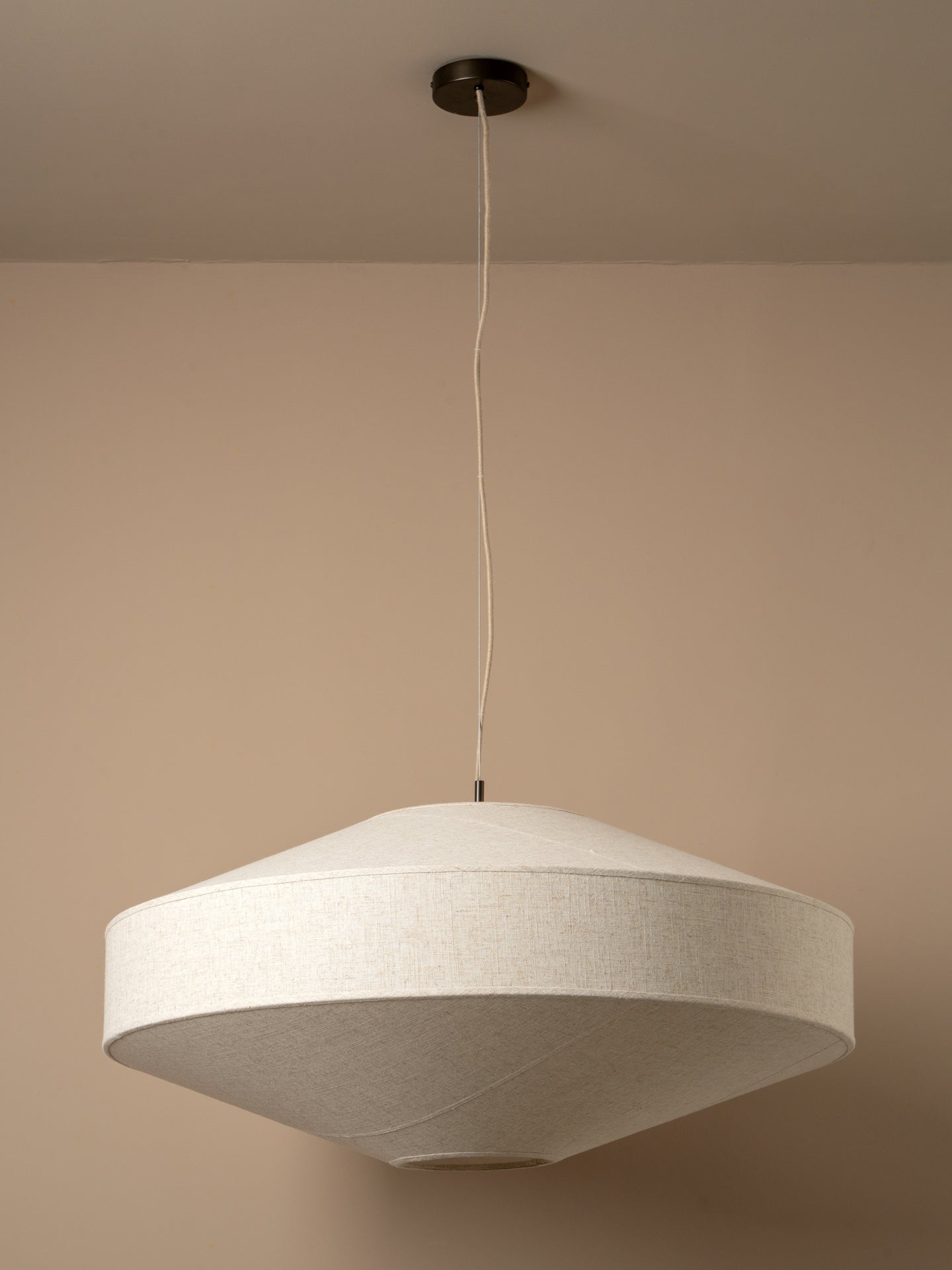 A modern pendant light with a wide, shallow conical shade made of textured beige fabric, suspended from a black ceiling canopy by a thin white cord against a warm beige wall.