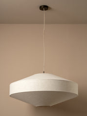 A modern pendant light with a wide, shallow conical shade made of textured beige fabric, suspended from a black ceiling canopy by a thin white cord against a warm beige wall.