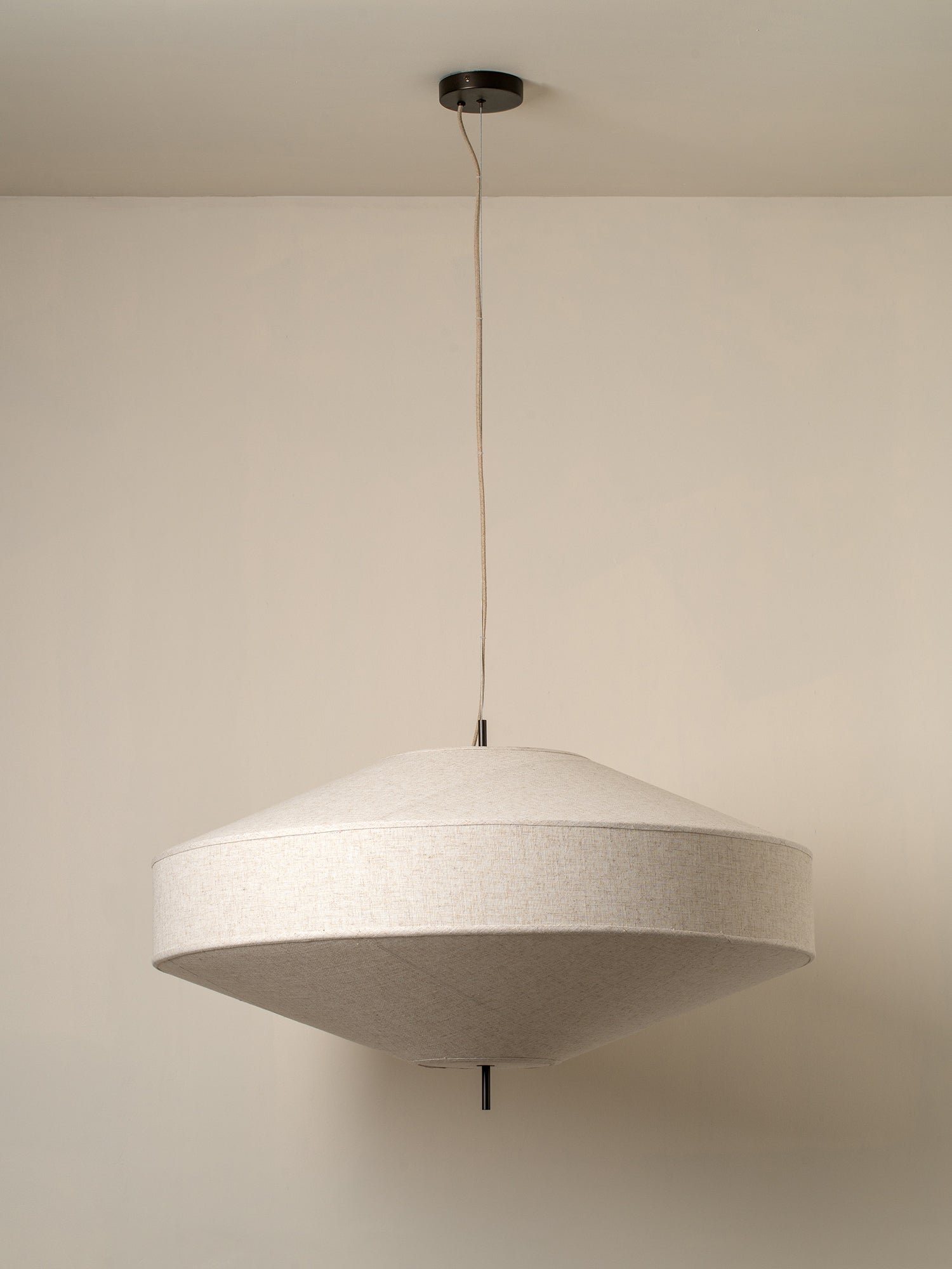 A modern pendant light with a wide, shallow conical shade made of textured off-white fabric, suspended by a thin cord from a black ceiling canopy against a plain beige wall.