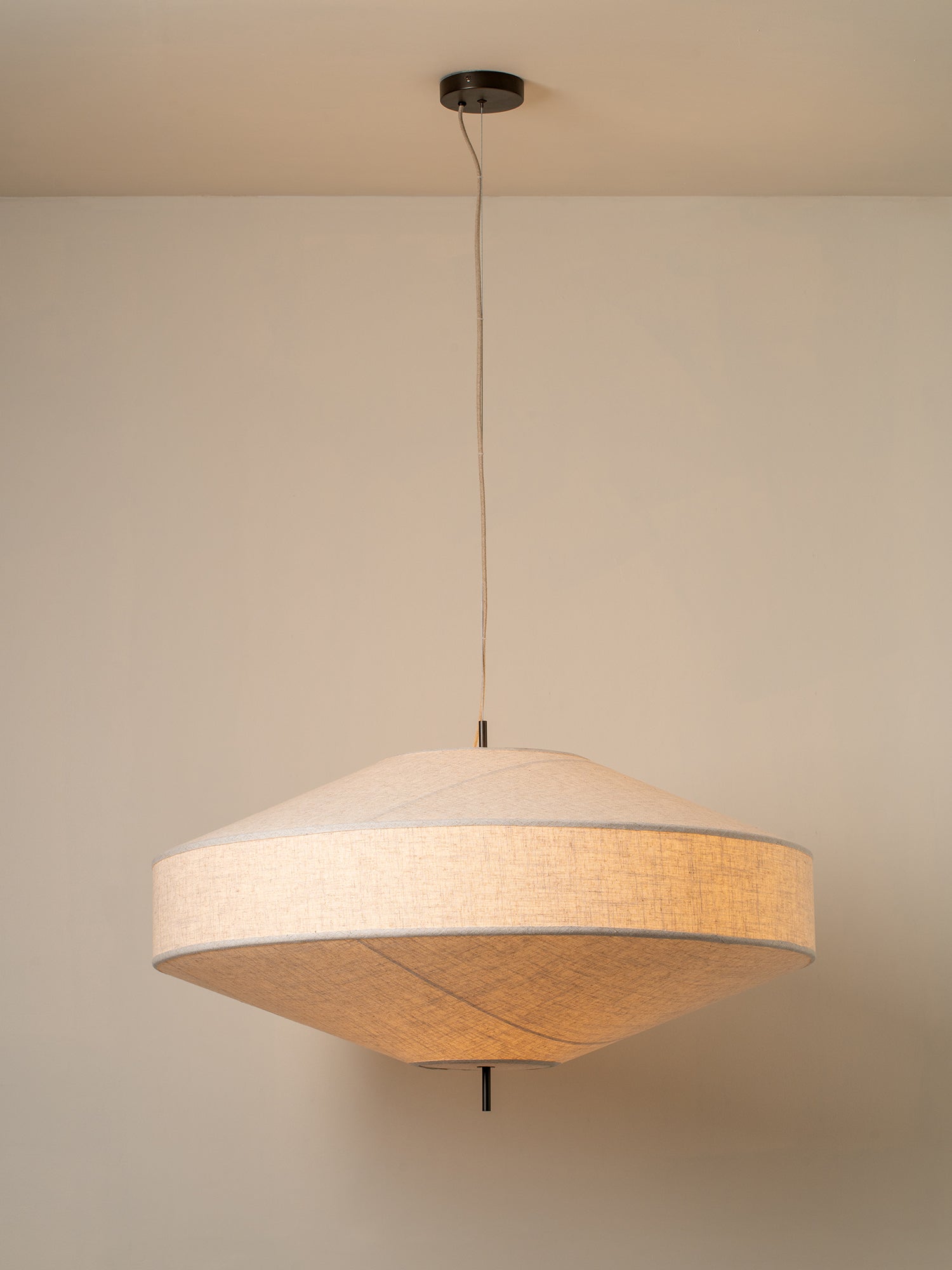 A modern pendant light with a textured beige fabric drum shade, suspended from a black ceiling canopy against a warm beige wall.