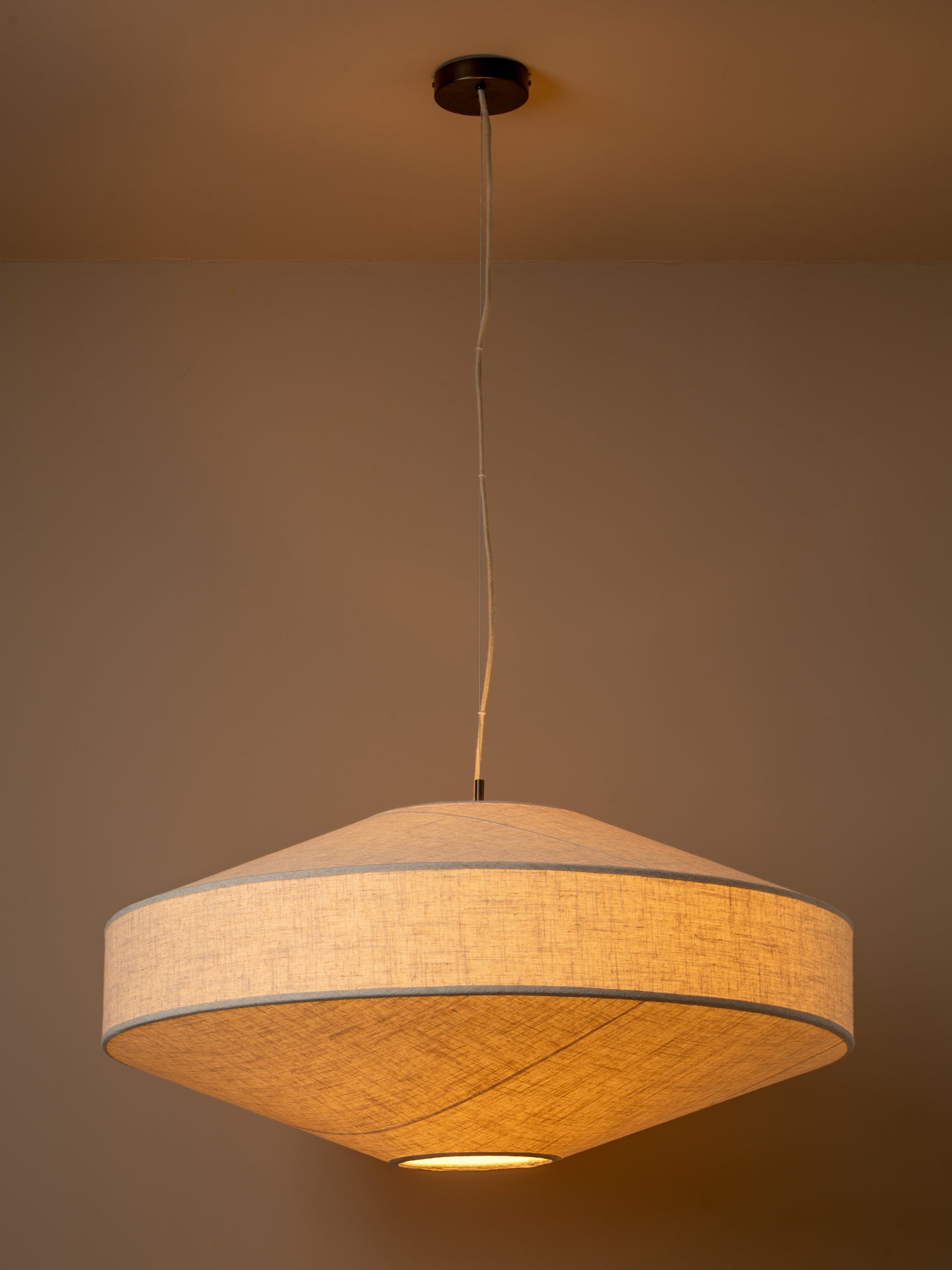 A modern pendant light with a wide, shallow conical shade made of textured beige fabric, suspended from a black ceiling canopy by a thin white cord against a warm beige wall. Light turned on.