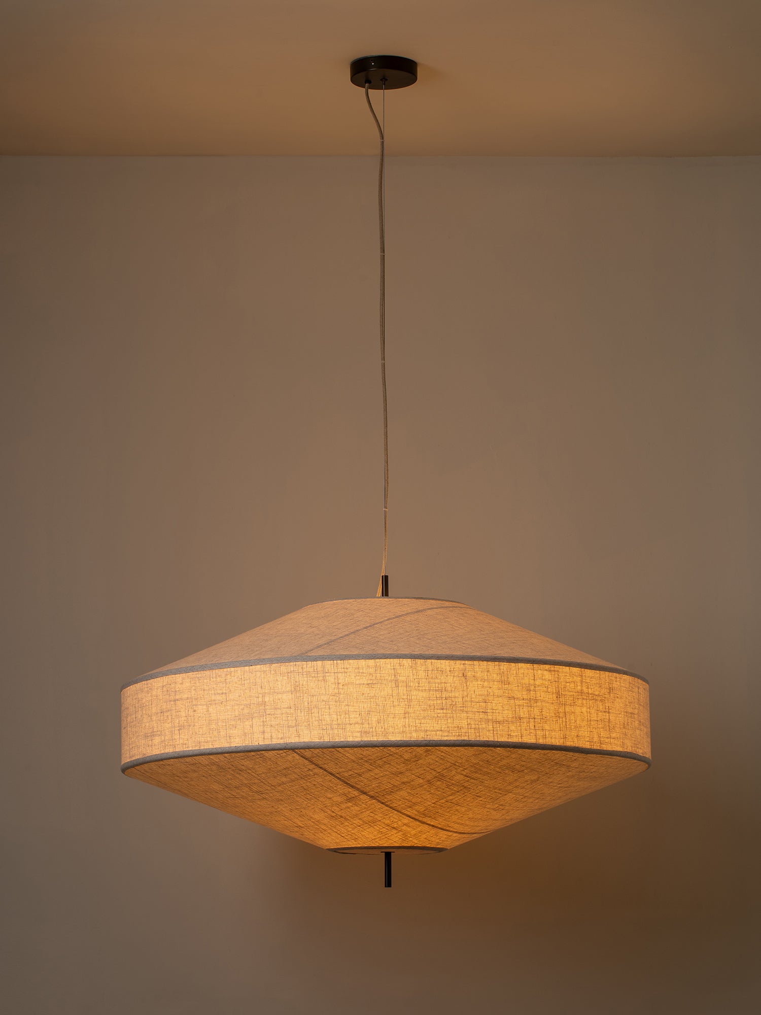 A modern pendant light with a textured beige linen shade, suspended by a thin black cord from a ceiling canopy against a neutral beige wall.
