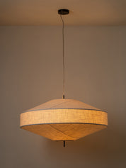 A modern pendant light with a textured beige linen shade, suspended by a thin black cord from a ceiling canopy against a neutral beige wall.