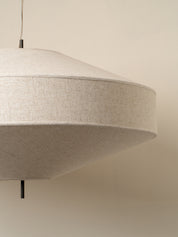 A close-up of a modern pendant light with a wide, conical shade made of textured beige linen fabric, suspended against a plain beige wall.