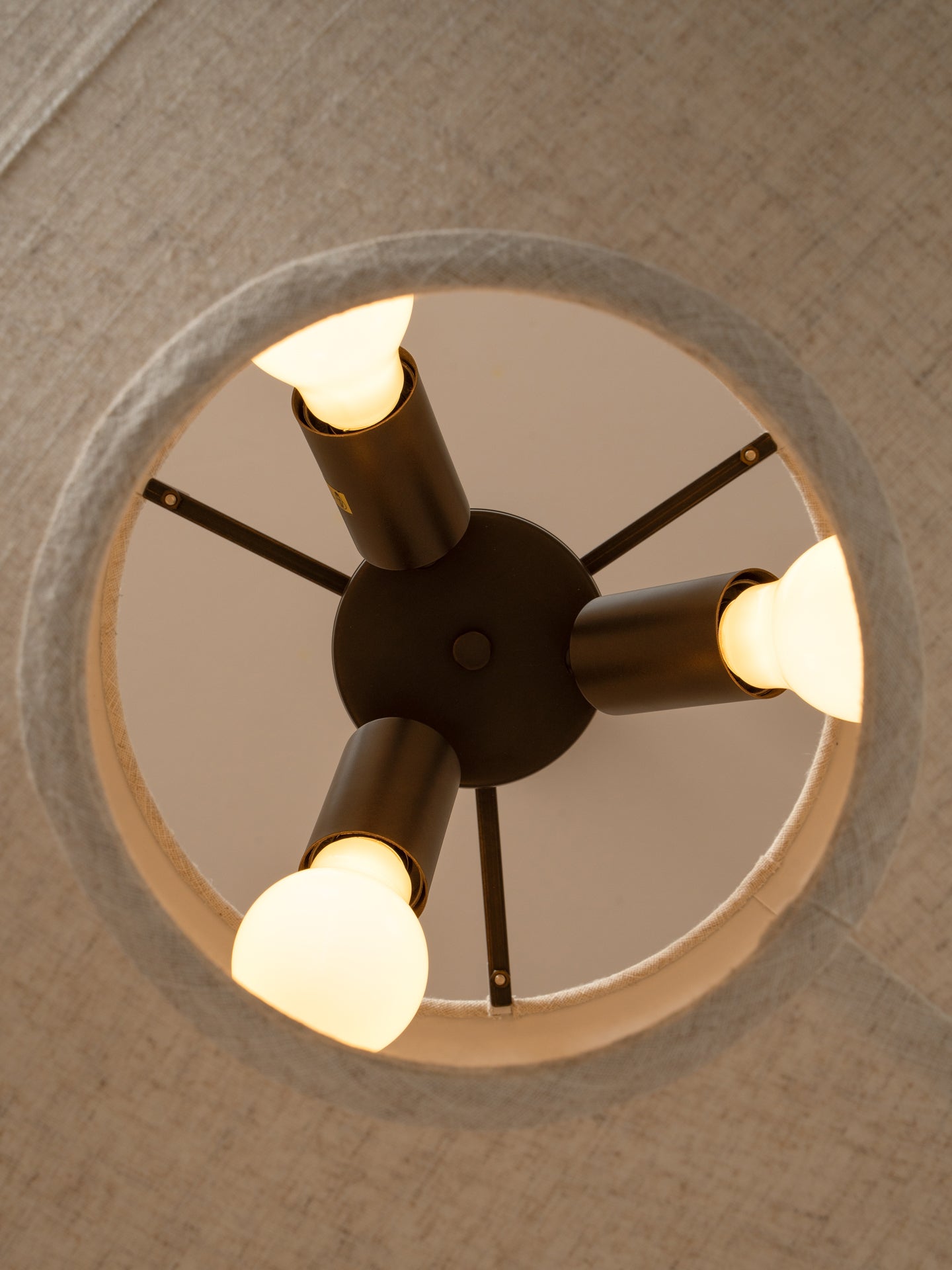 A modern four-light ceiling fixture with a dark central hub and cylindrical arms, each holding a glowing warm white bulb, shown installed on a textured beige ceiling.